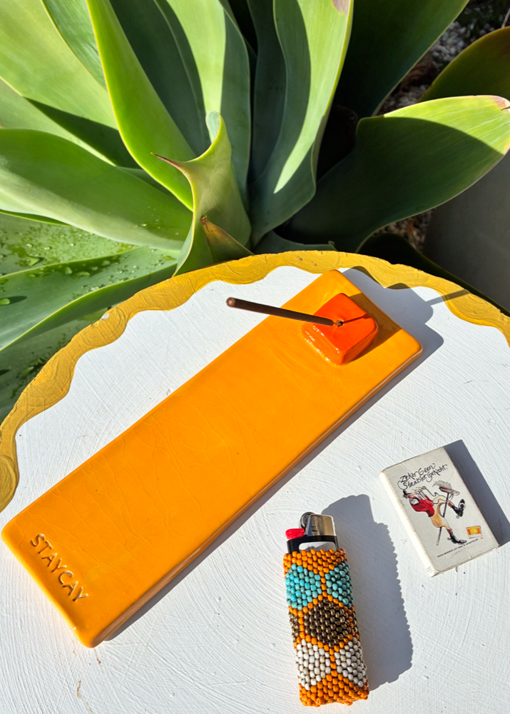 Handmade Kinga Csilla x Staycay ceramic incense holder in vibrant orange with red square detail, embossed with Staycay logo. Photographed outdoors with incense stick, matches and beaded lighter on white table, perfect for bohemian aromatherapy and home décor.