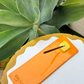 Bright orange Kinga Csilla x Staycay ceramic incense holder with a glossy yellow cube incense rest. Handmade artisan incense tray for meditation, rituals, and home styling. Photographed outdoors on a scalloped white and yellow table with lush green succulent backdrop.