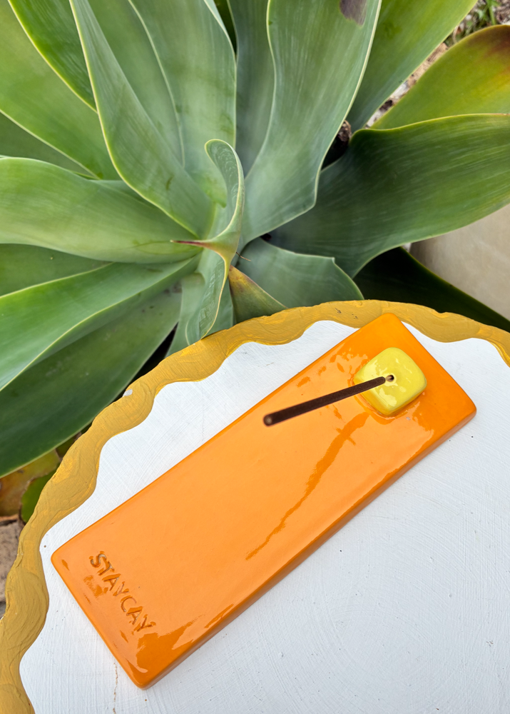 Bright orange Kinga Csilla x Staycay ceramic incense holder with a glossy yellow cube incense rest. Handmade artisan incense tray for meditation, rituals, and home styling. Photographed outdoors on a scalloped white and yellow table with lush green succulent backdrop.