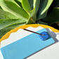 Sky blue Kinga Csilla x Staycay ceramic incense holder with a glossy deep blue cube incense rest. Handmade artisan incense tray for mindfulness, rituals, and home décor. Photographed outdoors on a scalloped white and yellow table with green succulent leaves in the background.