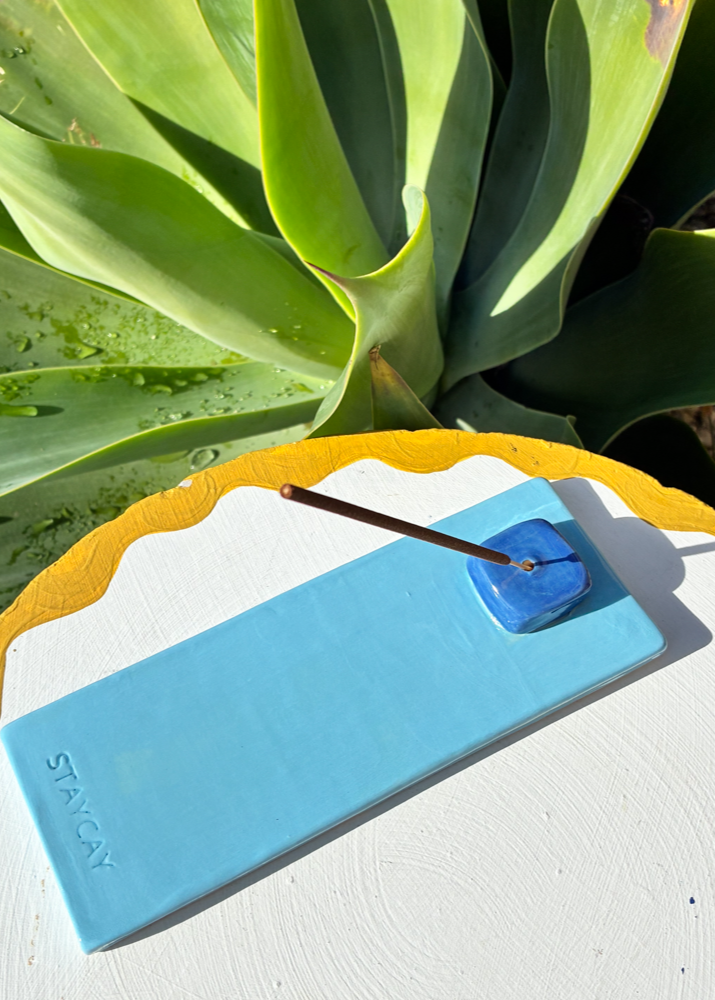 Sky blue Kinga Csilla x Staycay ceramic incense holder with a glossy deep blue cube incense rest. Handmade artisan incense tray for mindfulness, rituals, and home décor. Photographed outdoors on a scalloped white and yellow table with green succulent leaves in the background.