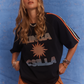 Close-up studio shot of model wearing Kinga Csilla Sunset Tee in black with sun logo and striped sleeve detail, styled with Bello Swirl Jessi Pants, against a blue backdrop