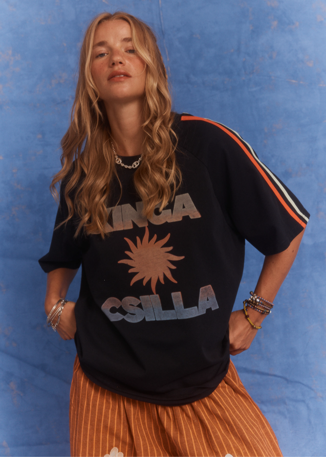 Close-up studio shot of model wearing Kinga Csilla Sunset Tee in black with sun logo and striped sleeve detail, styled with Bello Swirl Jessi Pants, against a blue backdrop