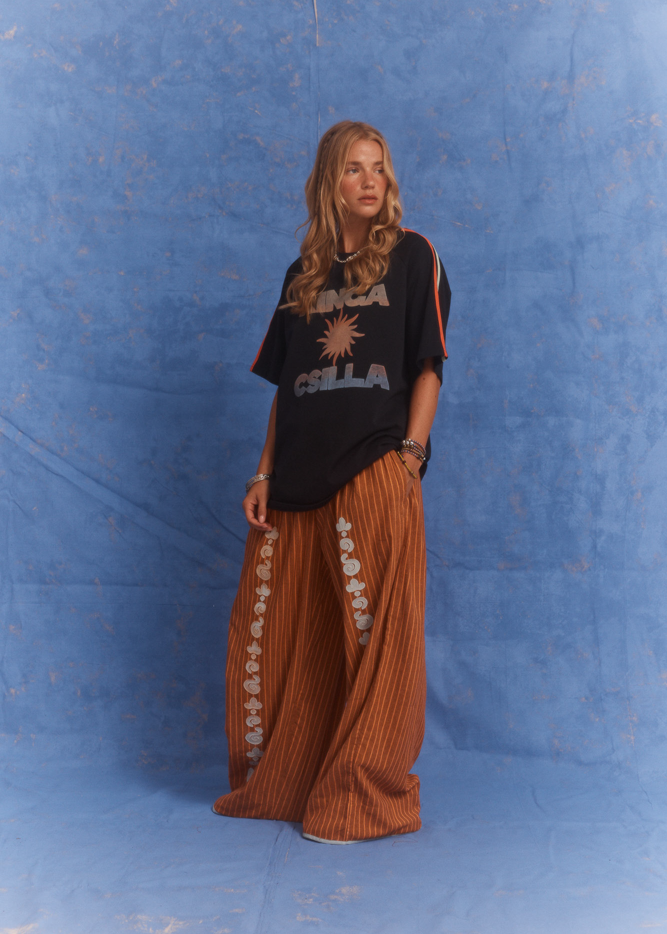 Studio shot of model wearing Kinga Csilla Sunset Tee in black with sun logo, paired with Bello Swirl Jessi Pants featuring floral embroidery, against a blue backdrop