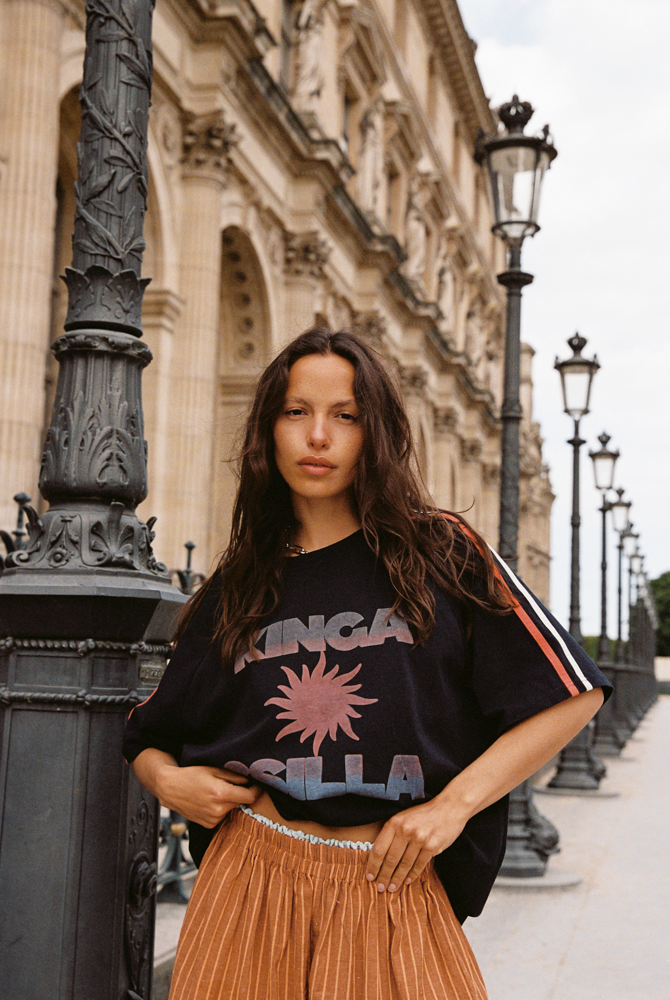 Model wearing Kinga Csilla Sunset Tee in black cotton with graphic sun print and striped sleeves, styled with Bello Swirl Jessi Pant in a street-style setting