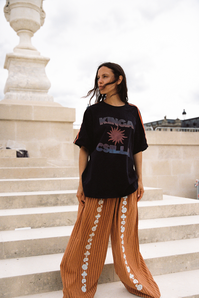 Model wearing Kinga Csilla Sunset Tee in black with sun logo, styled with Bello Swirl Jessi Pants featuring embroidered floral details, standing on stone steps in Paris