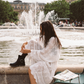 Woman wearing the Kinga Csilla Tottori Lace Harlow Maxi Dress in white embroidered cotton, styled with black ankle boots, sitting by a Paris fountain.