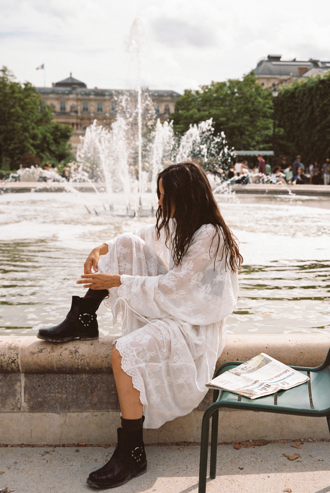 Woman wearing the Kinga Csilla Tottori Lace Harlow Maxi Dress in white embroidered cotton, styled with black ankle boots, sitting by a Paris fountain.