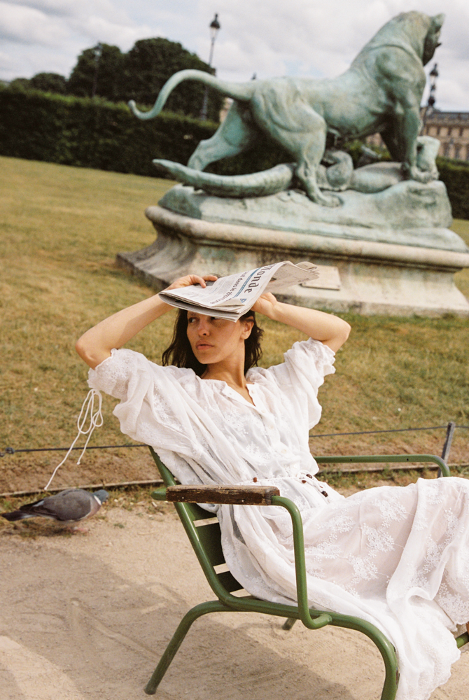 Model in the Kinga Csilla Tottori Lace Harlow Maxi Dress in white cotton lace, sitting on a Parisian garden chair holding a newspaper with a bronze statue in the background.
