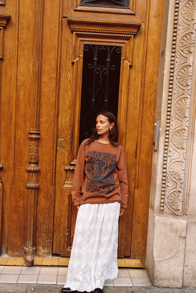 Woman wearing Kinga Csilla Tottori Lace Ode Skirt – white cotton lace maxi skirt styled with oversized brown Lino Roses long sleeve tee in Paris street style look