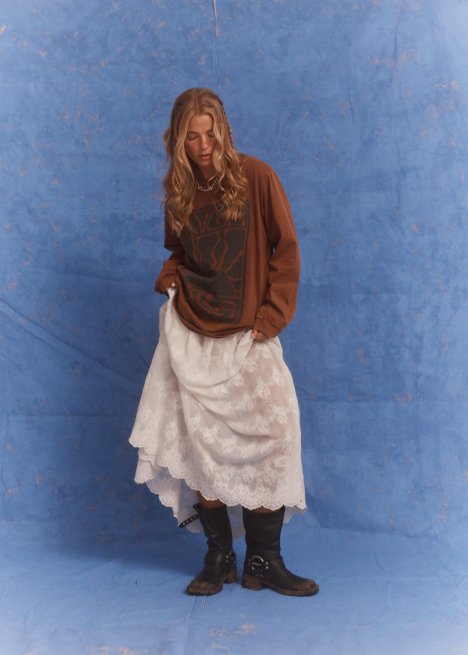 Model wearing Kinga Csilla Tottori Lace Ode Skirt, white lace maxi skirt styled with oversized brown Lino Roses Tee and black boots, studio photoshoot with blue backdrop