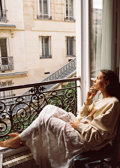 Model in Kinga Csilla Tottori Lace Ode Skirt, white lace maxi skirt styled with oversized graphic Shell Og Jumper, relaxing on Paris balcony with coffee cup