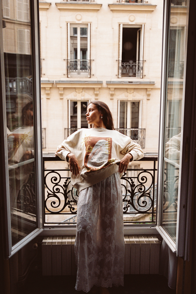 Model in Kinga Csilla Tottori Lace Ode Skirt, white cotton lace maxi skirt styled with Shell OG Jumper, standing on a Paris balcony editorial fashion look