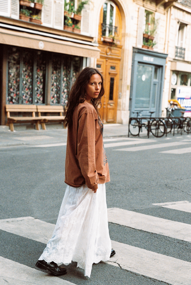 Model wearing Kinga Csilla Tottori Lace Ode Skirt, a white lace cotton maxi skirt, styled with oversized brown Lino Roses long sleeve tee in Paris street style look