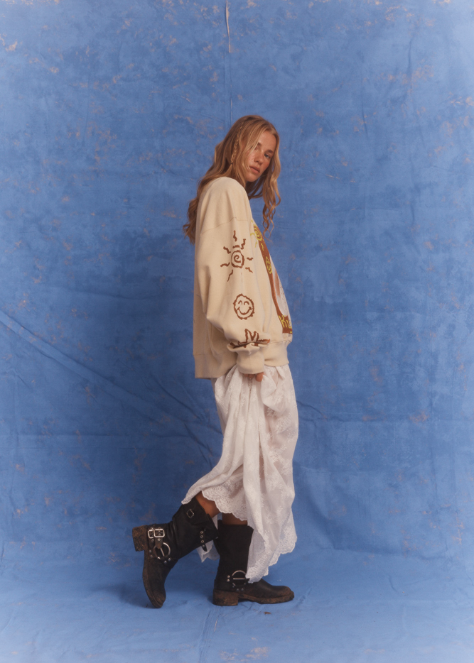 Side profile of model wearing Kinga Csilla Tottori Lace Ode Skirt, white lace maxi skirt styled with oversized beige Shell Og Jumper and black leather boots, studio photoshoot on blue backdrop