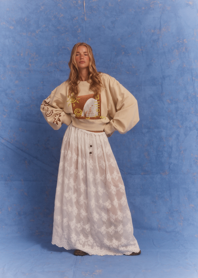 Model wearing Kinga Csilla Tottori Lace Ode Skirt, white lace maxi skirt styled with oversized beige Shell OG Jumper, studio photoshoot on blue backdrop