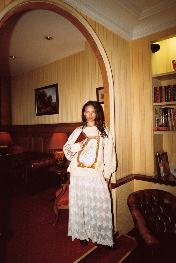 Model wearing Kinga Csilla Tottori Lace Ode Skirt, white lace maxi skirt styled with Shell OG Jumper, standing in vintage library-inspired interior look