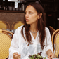 Model wearing the Kinga Csilla Tottori Lace Pia Mini in white cotton lace embroidery, dining outdoors at a Paris café, styled with silver chain necklace.