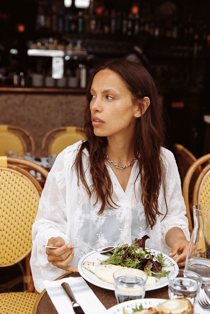 Model wearing the Kinga Csilla Tottori Lace Pia Mini in white cotton lace embroidery, dining outdoors at a Paris café, styled with silver chain necklace.