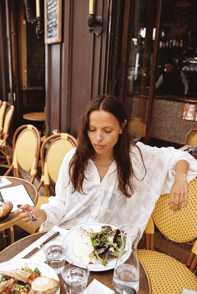 Woman in Kinga Csilla Tottori Lace Pia Mini white embroidered cotton dress sitting at a Paris café table with salad and omelette, relaxed French lifestyle outfit inspiration.