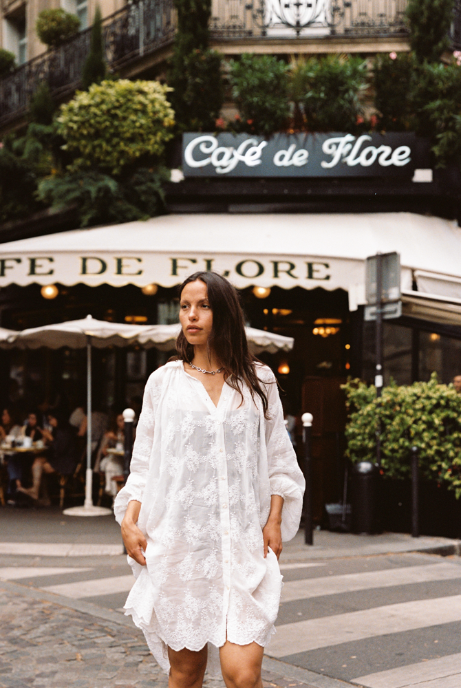 Woman wearing the Kinga Csilla Tottori Lace Pia Mini in white lace, styled with scalloped hem and embroidery, photographed in Paris street style outside Café de Flore.
