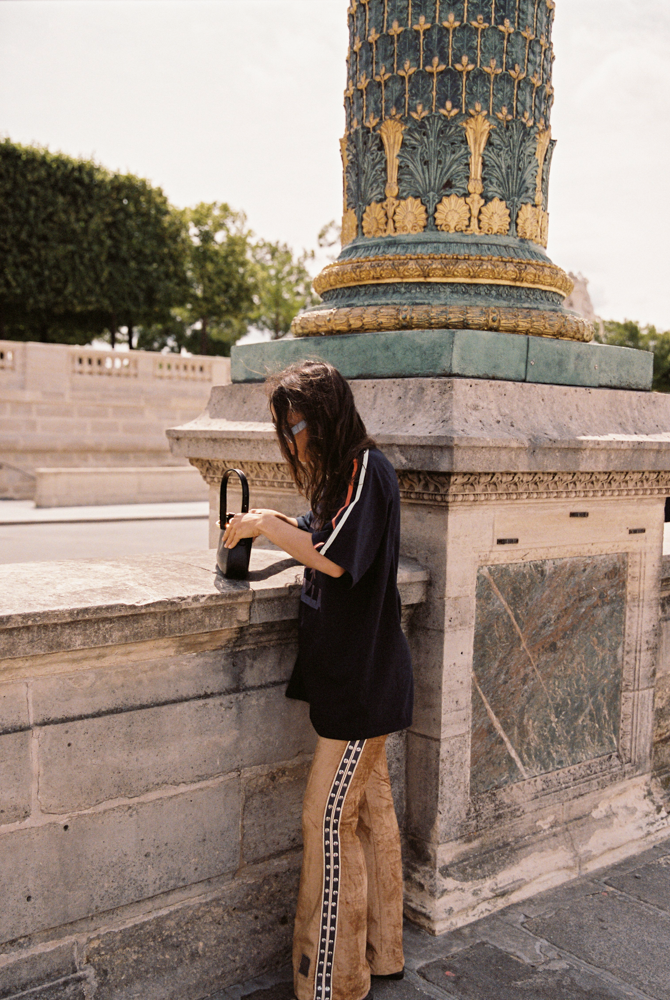 Model in Kinga Csilla Twilight Velvet Flare Pant in golden velvet with KC logo stripe detail, styled with oversized Kinga Csilla Sunset tee and mini shoulder bag, photographed against Paris architecture.