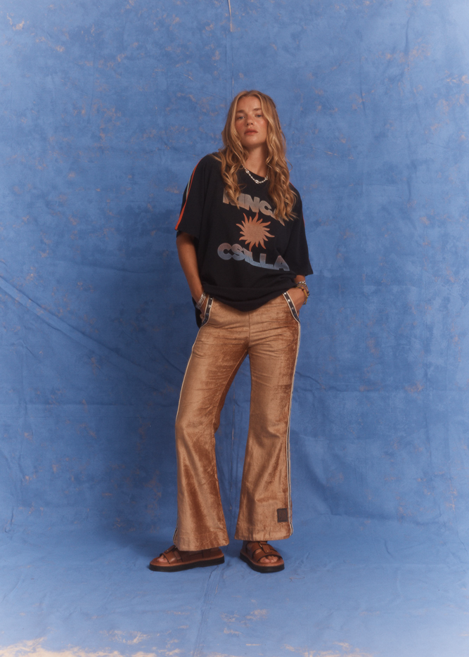 Model wearing Kinga Csilla Twilight Velvet Flare Pant in golden velvet with KC logo stripe, styled with oversized Kinga Csilla Sunset tee, photographed against a blue studio backdrop.