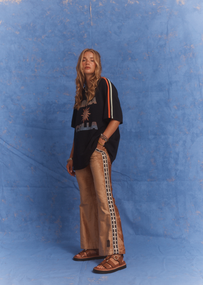 Model wearing Kinga Csilla Twilight Velvet Flare Pant in golden velvet with black logo side stripe detail, styled with oversized Kinga Csilla Sunset tee, photographed against a blue studio backdrop.