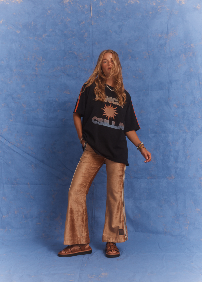 Model in Kinga Csilla Twilight Velvet Flare Pant in golden velvet with KC logo side stripe, styled with oversized Kinga Csilla Sunset tee, photographed against a textured blue backdrop.