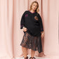 A woman models a black oversized V-neck tunic top with purple trim and a floral graphic on the chest, paired with a sheer black lace midi skirt and dark brown strappy sandals, set against a pink draped fabric backdrop.