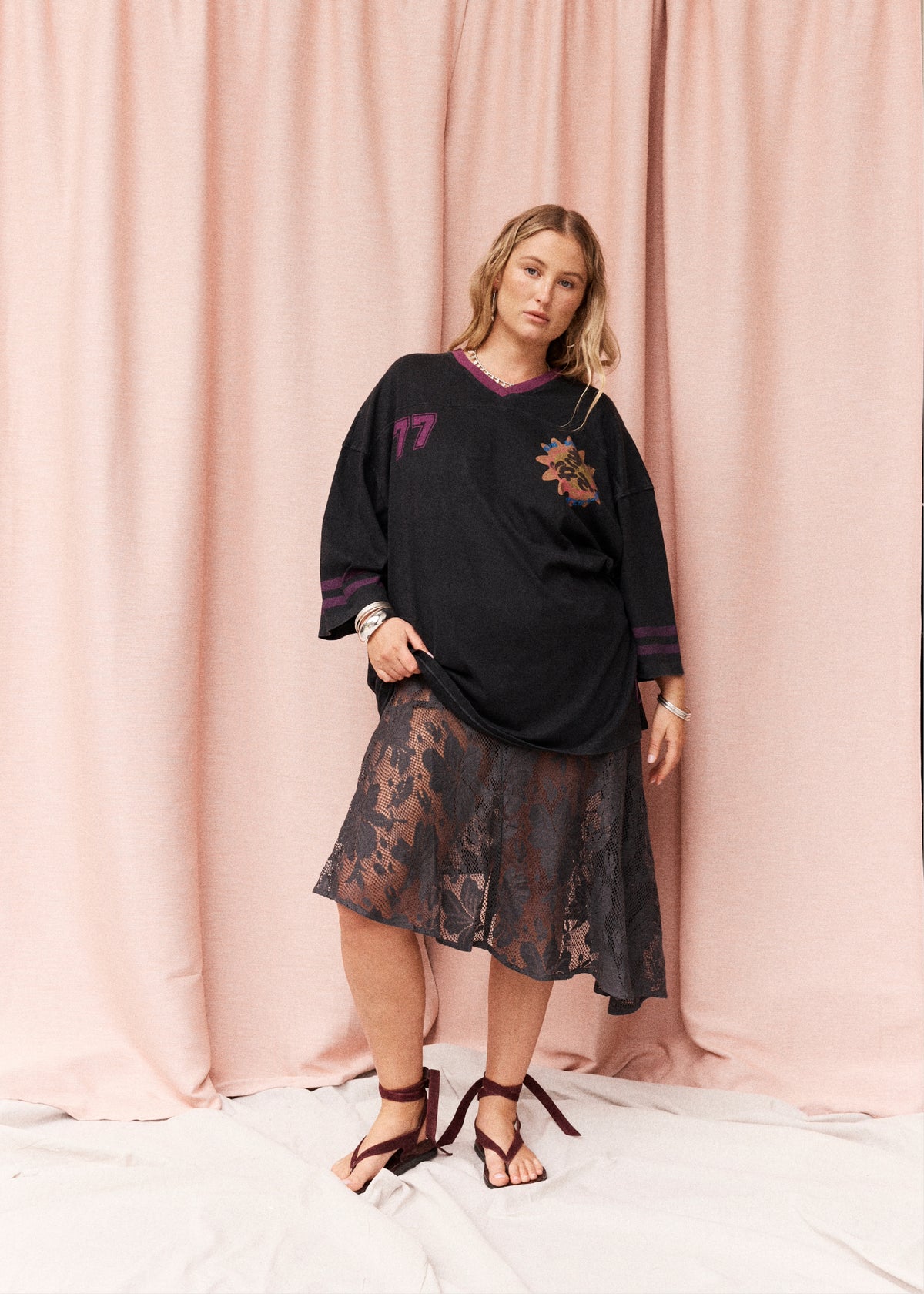 A woman models a black oversized V-neck tunic top with purple trim and a floral graphic on the chest, paired with a sheer black lace midi skirt and dark brown strappy sandals, set against a pink draped fabric backdrop.