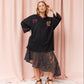 A woman models an oversized black vintage-style sports jersey top with purple trim and a floral graphic, paired with a sheer black lace skirt and dark wedge sandals, set against a pink draped fabric backdrop.