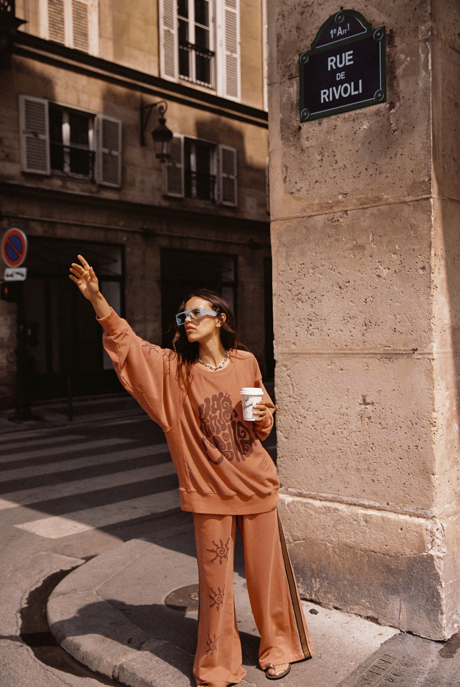 Woman in an orange tracksuit set standing on a street corner in Paris, holding a coffee cup.