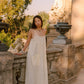 A woman wearing a cream sleeveless maxi dress and black sandals stands on an outdoor stone terrace with statues and trees in the background.