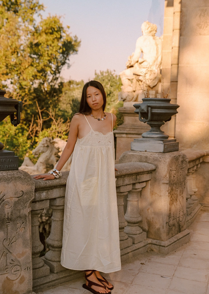 A woman wearing a cream sleeveless maxi dress and black sandals stands on an outdoor stone terrace with statues and trees in the background.