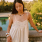 A woman wearing a cream lace-trimmed spaghetti-strap dress and silver bracelets stands outdoors by a stone railing with a pool and greenery in the background.