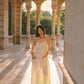 A woman stands in a sunlit stone colonnade wearing a flowing cream maxi dress and brown sandals, with columns and greenery visible in the background.