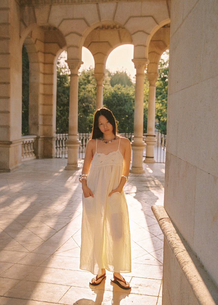 A woman stands in a sunlit stone colonnade wearing a flowing cream maxi dress and brown sandals, with columns and greenery visible in the background.