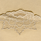 A close-up of a cream embroidered lace applique with a floral pattern, laid flat on a matching beige textured background.