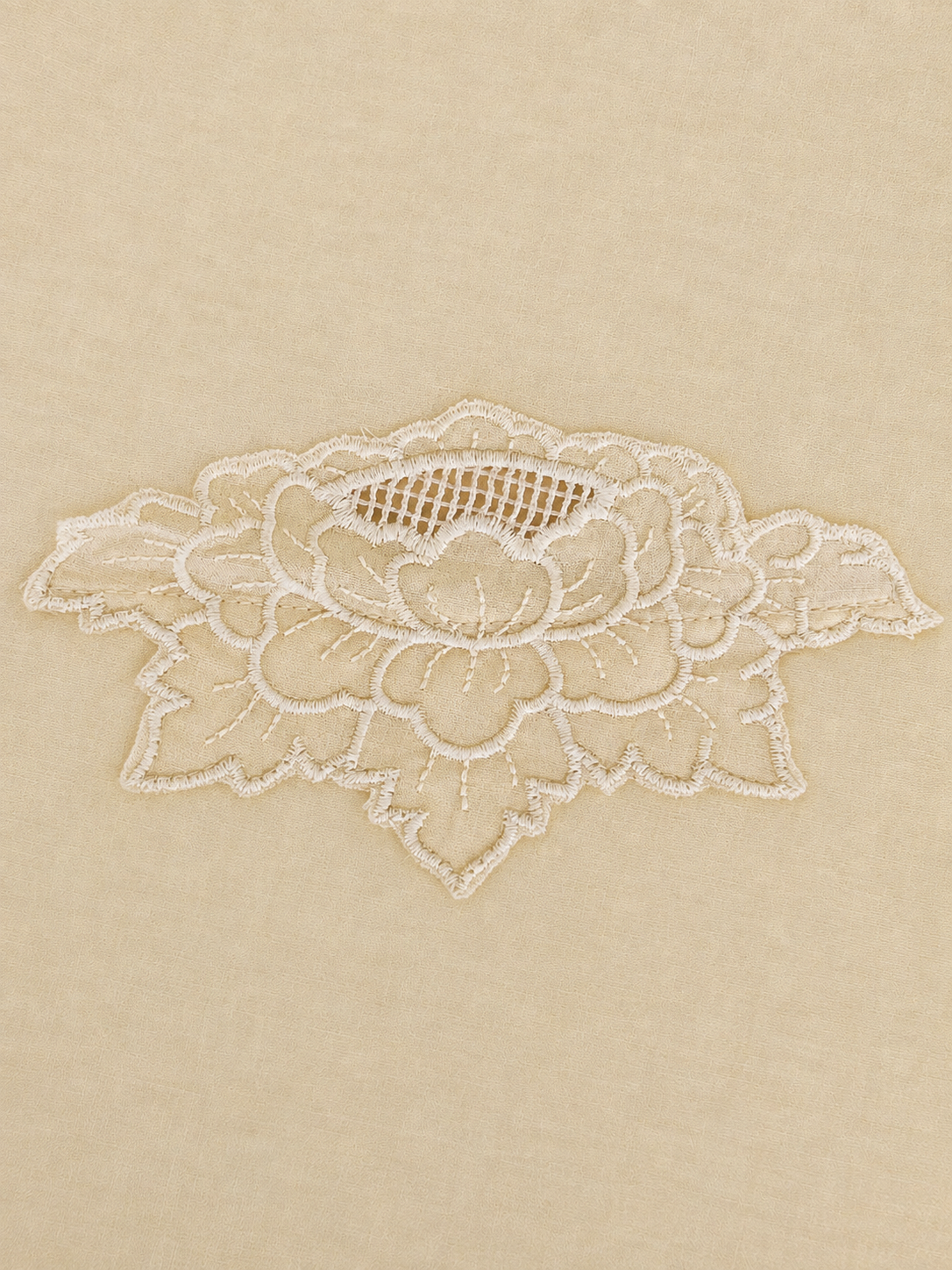 A close-up of a cream colored fabric surface featuring a delicate white floral lace applique patch.