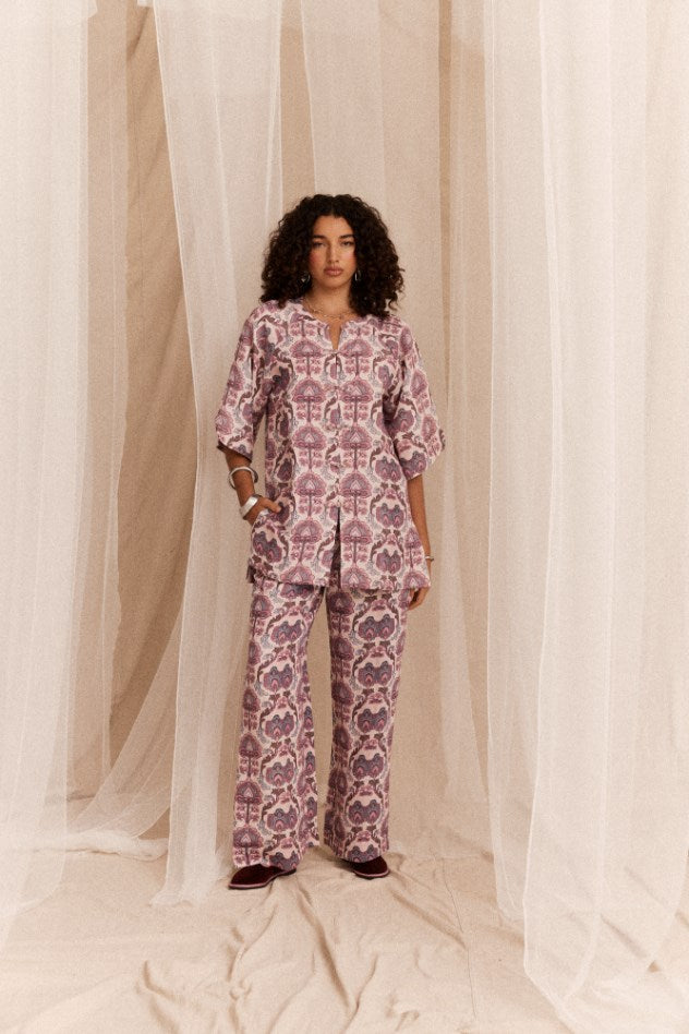 Woman wearing a pink patterned shirt and pant set standing in front of sheer curtains