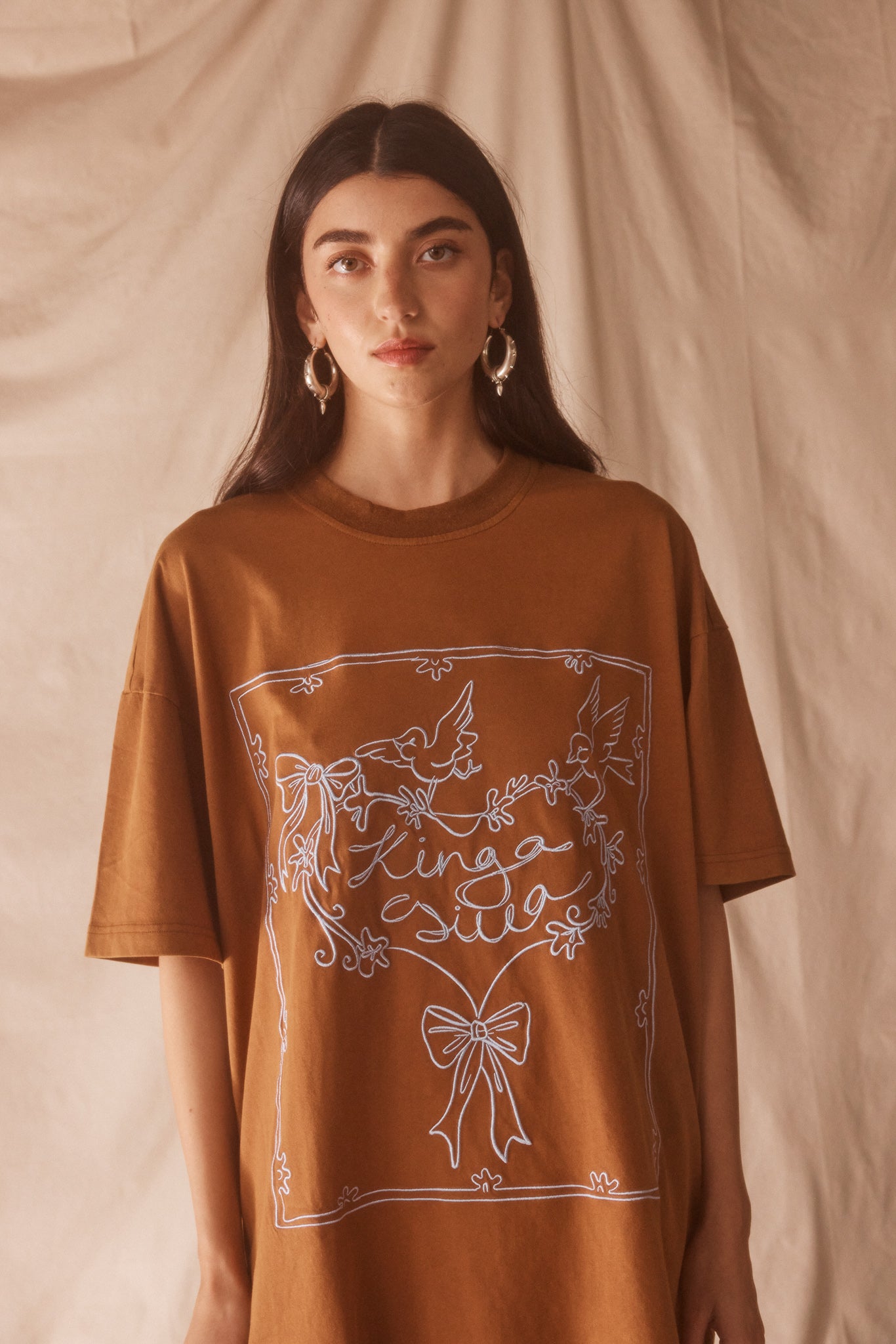 Woman wearing a brown oversized t-shirt with a baby blue graphic design against a beige background