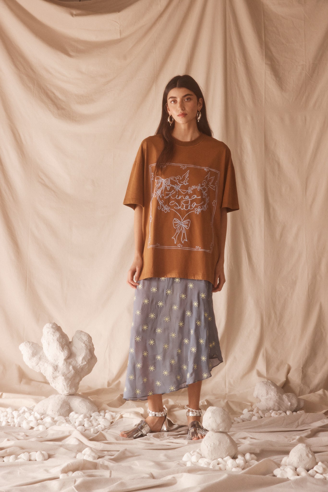 Woman wearing a oversized brown t-shirt with a baby blue graphic design against a beige curtain background