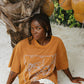 Woman in a brown oversized t-shirt sitting under a palm tree with coconuts.