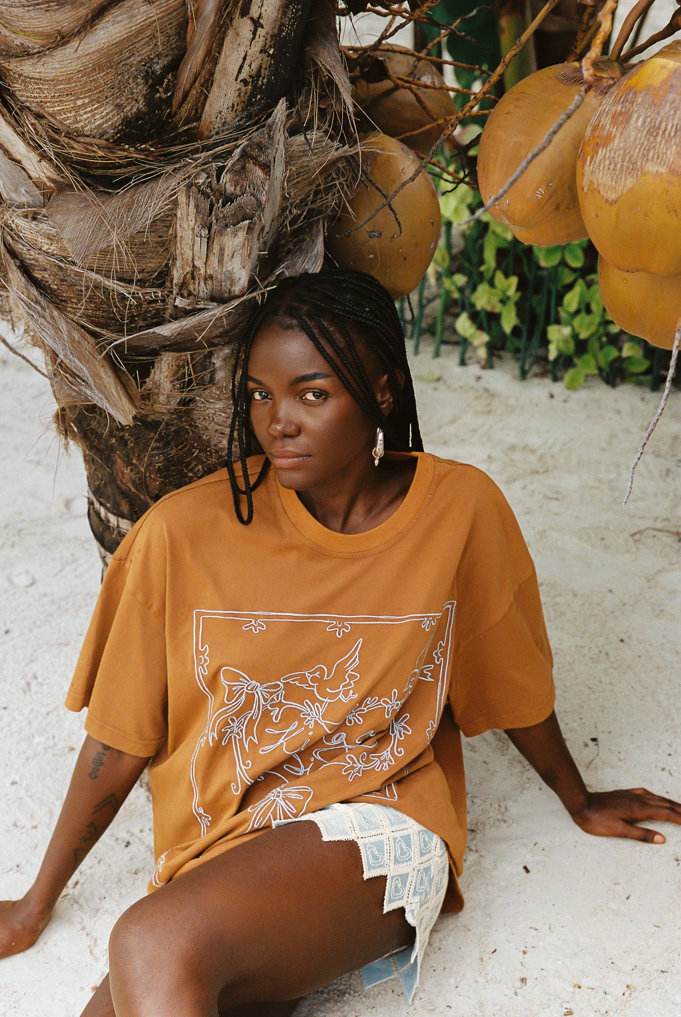 Woman in a brown oversized t-shirt sitting under a palm tree with coconuts.