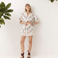 A woman stands against a white wall wearing a white short-sleeve button-front dress with colorful floral embroidery and a lace hem, paired with black strappy sandals. Green leaves hang from the top corners of the frame.