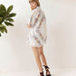 A woman wearing a white embroidered kimono-style cover-up with colorful floral patterns and a scalloped hem, paired with dark brown strappy sandals, standing against a white wall with green plants in the background.