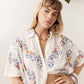 A woman wearing a white short-sleeve button-up shirt with colorful floral embroidery, posed against a white wall with green leaves hanging from above.