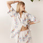 A woman modeling a white two-piece bohemian lounge set featuring a short-sleeve button-up shirt and matching shorts, both decorated with colorful floral prints and white lace trim, set against a neutral background with a green plant leaf in the corner.