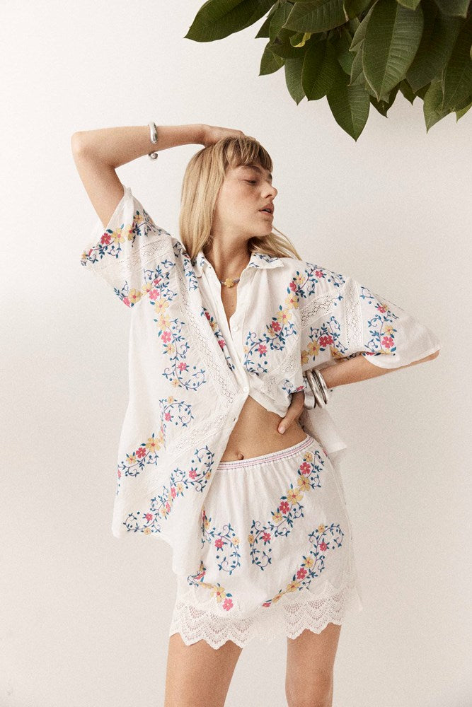 A woman modeling a white two-piece bohemian lounge set featuring a short-sleeve button-up shirt and matching shorts, both decorated with colorful floral prints and white lace trim, set against a neutral background with a green plant leaf in the corner.